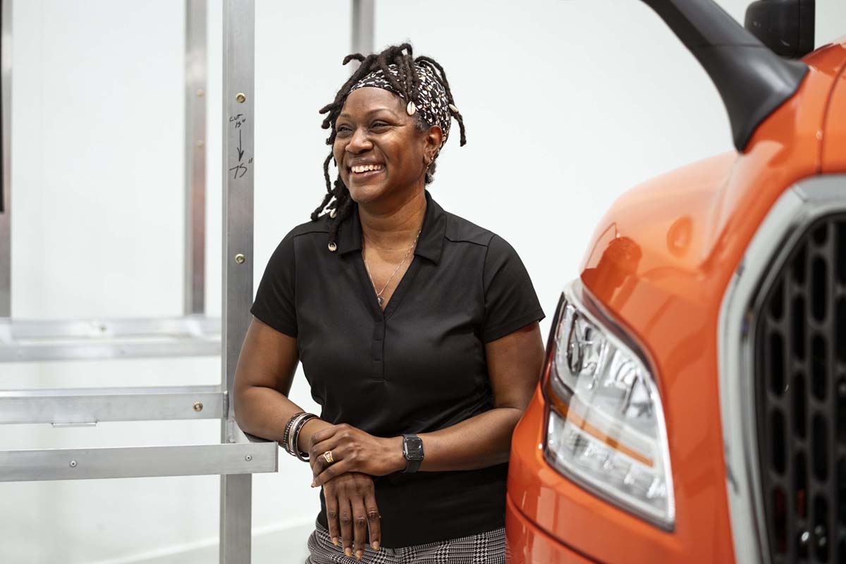 Female African American factory worker, laughing, leaning against a work bench. Next to and orange International MV.